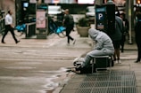 A person in a grey hoodie is sitting on a chair near a crosswalk, appearing to be resting or possibly homeless. They are surrounded by pedestrians walking by, some carrying bags. Bicycles are docked at a nearby bike-sharing station. Urban street elements such as a parking sign and wet pavement are visible.