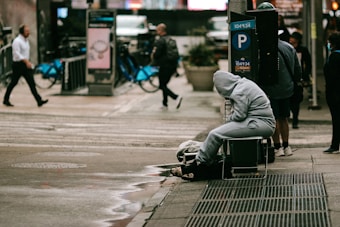 A person in a grey hoodie is sitting on a chair near a crosswalk, appearing to be resting or possibly homeless. They are surrounded by pedestrians walking by, some carrying bags. Bicycles are docked at a nearby bike-sharing station. Urban street elements such as a parking sign and wet pavement are visible.