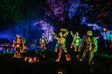 Artists carving eerie pumpkin sculptures illuminated by green and purple neon lights.