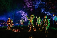 A collection of intricately carved pumpkins illuminated from within, arranged to resemble skeleton figures in a dark outdoor setting. The scene is enhanced by eerie, colorful lighting that casts shadows and highlights the carved details, creating a festive Halloween atmosphere.
