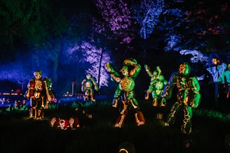A collection of intricately carved pumpkins illuminated from within, arranged to resemble skeleton figures in a dark outdoor setting. The scene is enhanced by eerie, colorful lighting that casts shadows and highlights the carved details, creating a festive Halloween atmosphere.