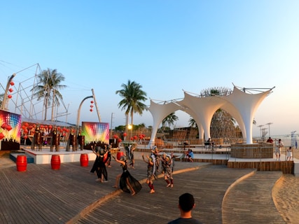 A vibrant outdoor performance featuring dancers in costumes, set against a backdrop of large, decorative canvases with multicolored lights. Palm trees and an architectural installation made of woven materials are present. The atmosphere is lively with people gathered around an open, wooden platform.