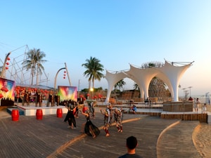 A vibrant outdoor performance featuring dancers in costumes, set against a backdrop of large, decorative canvases with multicolored lights. Palm trees and an architectural installation made of woven materials are present. The atmosphere is lively with people gathered around an open, wooden platform.