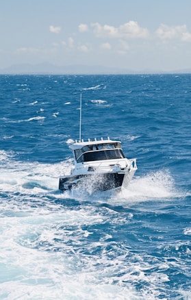A small white motorboat speeds across the ocean, creating splashes and waves behind it. The sea is a vibrant blue, and the sky is clear with a few scattered white clouds.