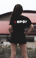 A person with long hair is standing outdoors, wearing a black t-shirt with the text 'G SPOT' on the back. The background includes a building with a red roof, and there is foliage in the foreground.