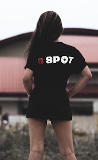 A person with long hair is standing outdoors, wearing a black t-shirt with the text 'G SPOT' on the back. The background includes a building with a red roof, and there is foliage in the foreground.