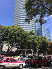 A modern, cylindrical skyscraper with reflective glass windows rises behind lush green trees. Several cars, including a pink and white taxi with the sign 'CDMX TAXI', are seen in the foreground, indicating a busy urban street setting.