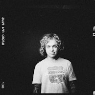 A monochrome image featuring a person with curly hair wearing a t-shirt that has a logo or graphic design on it. The background is dark and the person is centered, looking directly at the camera.