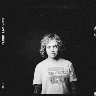 A monochrome image featuring a person with curly hair wearing a t-shirt that has a logo or graphic design on it. The background is dark and the person is centered, looking directly at the camera.