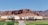 Several covered wagons are positioned on a grass and dirt area with a backdrop of striking red rock cliffs under a clear blue sky. The wagons are arranged as part of a campground or lodging setup, with wooden decking and a group of red chairs visible in the center.