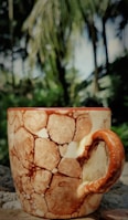 A ceramic mug with a brown and cream marbled pattern sits on a stone surface. The intricate design on the mug resembles natural stone textures. In the blurred background, there are lush green trees and vegetation, with sunlight filtering through the leaves.