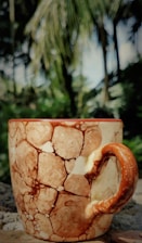 A ceramic mug with a brown and cream marbled pattern sits on a stone surface. The intricate design on the mug resembles natural stone textures. In the blurred background, there are lush green trees and vegetation, with sunlight filtering through the leaves.