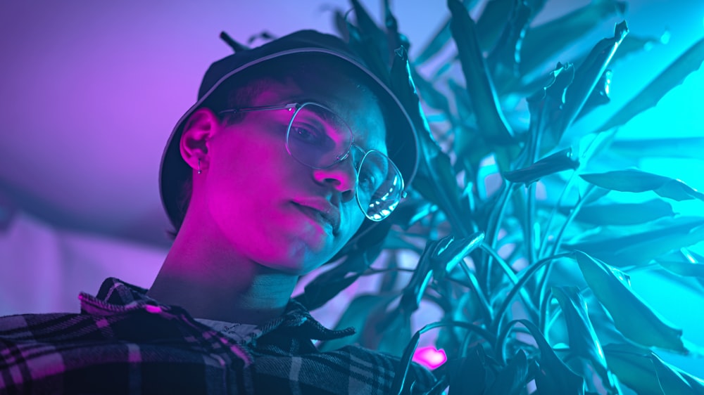 a man wearing glasses and a hat standing next to a plant