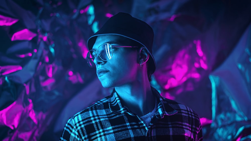 a man wearing glasses and a hat standing in front of a purple background