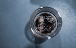 A top-down view of a glass filled with a dark carbonated drink and ice cubes. The liquid appears to have small bubbles on the surface. The background is a textured gray surface.