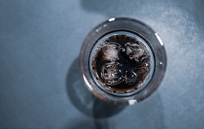 A top-down view of a glass filled with a dark carbonated drink and ice cubes. The liquid appears to have small bubbles on the surface. The background is a textured gray surface.