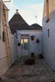 A narrow stone pathway leads to a small house with conical rooftops typical of trulli architecture. The walls are painted white, and there are several potted plants with red flowers placed on the ground and mounted on walls. There's a ladder on the left side and an antenna above the roof.