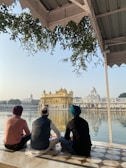 Three individuals sit cross-legged beside a reflective pool, gazing at a majestic, gold-covered temple across the water. The temple is surrounded by a marble walkway, and lush greenery hangs from an overhang above.