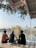 Three individuals sit cross-legged beside a reflective pool, gazing at a majestic, gold-covered temple across the water. The temple is surrounded by a marble walkway, and lush greenery hangs from an overhang above.