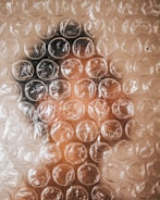 A face is seen through a sheet of clear, textured bubble wrap. The bubble wrap creates a distorted effect, obscuring details while allowing hints of the face to be visible. Light reflections are visible on the surface of the bubbles, adding to the texture.