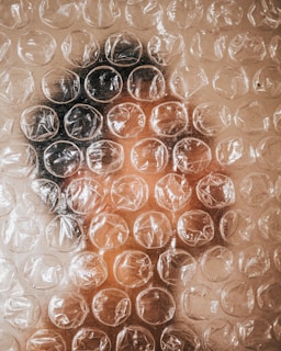 A face is seen through a sheet of clear, textured bubble wrap. The bubble wrap creates a distorted effect, obscuring details while allowing hints of the face to be visible. Light reflections are visible on the surface of the bubbles, adding to the texture.
