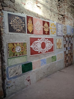A wall displays a variety of decorative tiles arranged in a grid-like pattern. Each tile features intricate designs, including floral motifs and geometric patterns, in colors like red, yellow, brown, and blue. The wall itself is made of exposed brick and concrete, giving a rustic appearance.