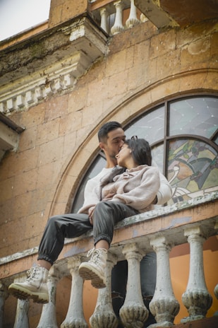 A couple sits intimately on the railing of an old, rustic building. The man embraces the woman as they share a tender moment, with an intricate stained glass window in the background. The building features weathered stone and delicate architectural details, creating a nostalgic atmosphere.