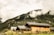 A series of wooden cabins with sloped roofs are nestled at the base of a lush, forested hillside. Thick clouds partially cover the hill, creating a misty atmosphere. The foreground is filled with tall grasses and small flowering plants, adding a rustic charm to the scene.