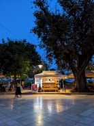A brightly lit kiosk is set in a park-like area during evening time. The kiosk displays snacks and drinks with a glowing light, making it an inviting spot against the darkening sky. People are casually walking by and sitting at nearby tables under the dim light of surrounding trees.
