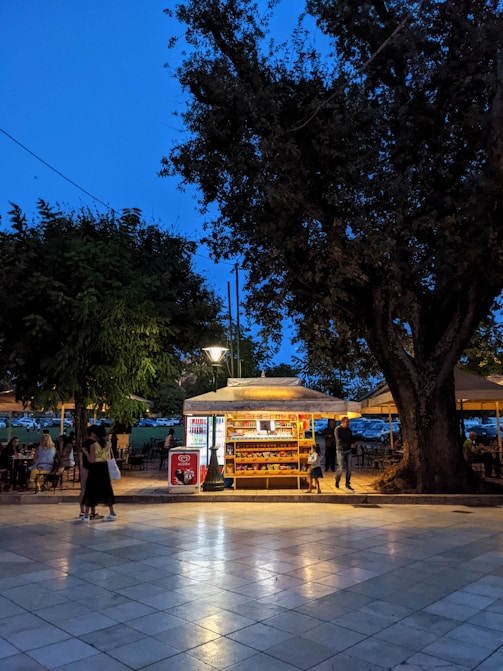 A brightly lit kiosk is set in a park-like area during evening time. The kiosk displays snacks and drinks with a glowing light, making it an inviting spot against the darkening sky. People are casually walking by and sitting at nearby tables under the dim light of surrounding trees.