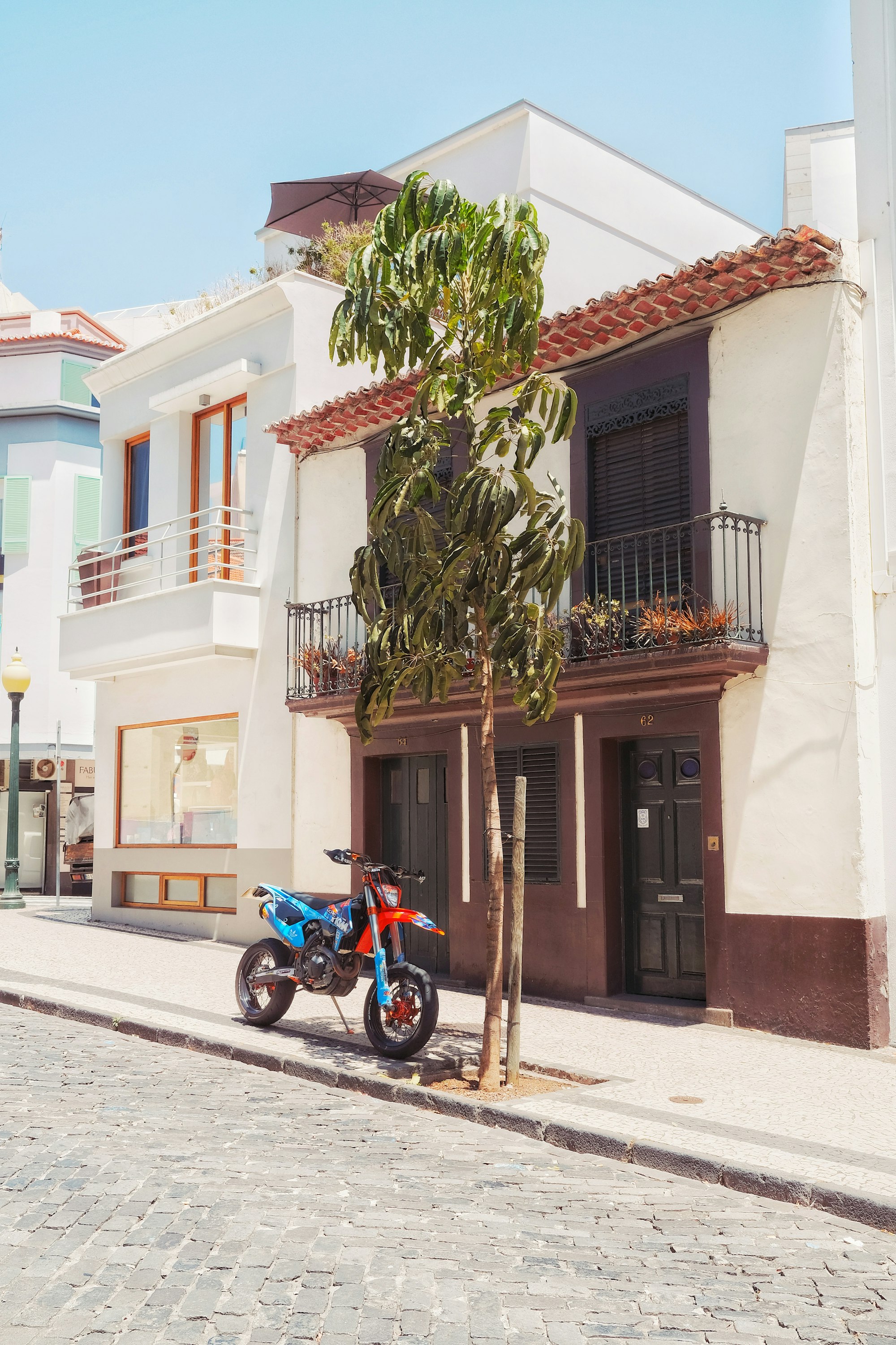 A blue and orange motorcycle is seen beneath a green tree in summer on a side street in Funchal, Madeira