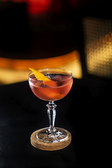 A cocktail glass filled with an amber-colored liquid, garnished with a twist of orange peel, is placed on a cork coaster against a dark, dimly lit background.