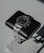 A luxury wristwatch with a black dial and a stainless steel band is positioned on a cushioned black stand. The stand is set on a marble base, creating a sophisticated and elegant presentation.