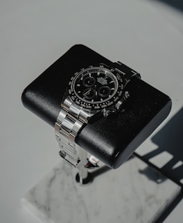 A luxury wristwatch with a black dial and a stainless steel band is positioned on a cushioned black stand. The stand is set on a marble base, creating a sophisticated and elegant presentation.