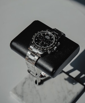 A luxury wristwatch with a black dial and a stainless steel band is positioned on a cushioned black stand. The stand is set on a marble base, creating a sophisticated and elegant presentation.