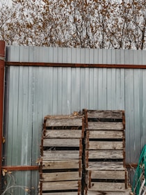 A tall metal fence with a rustic, weathered appearance, supported by rust-colored metal brackets. In front of the fence, there are two wooden pallets leaning against it, and a green hose is coiled nearby. Behind the fence, bare branches with scattered brown leaves reach into the cloudy sky.