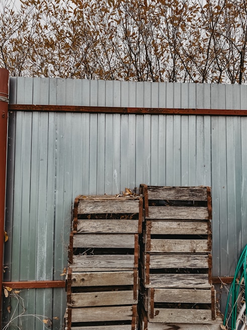 A tall metal fence with a rustic, weathered appearance, supported by rust-colored metal brackets. In front of the fence, there are two wooden pallets leaning against it, and a green hose is coiled nearby. Behind the fence, bare branches with scattered brown leaves reach into the cloudy sky.