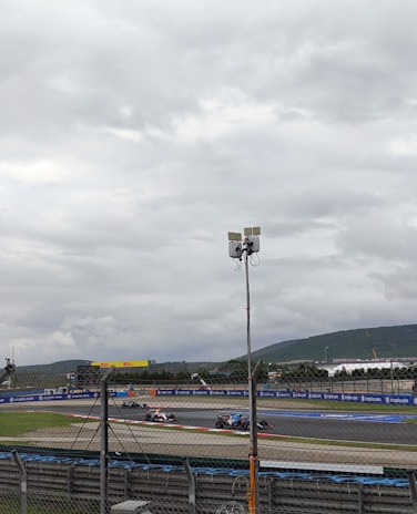 A race track with several race cars speeding along a curve. The sky is overcast with thick clouds. There are hills visible in the background and the track is enclosed by fences and advertising boards. A tall pole with cameras or sensors is positioned prominently in the foreground.