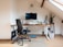 A modern, organized home office setup in an attic space features a sleek desk with a computer monitor, keyboard, and mouse. Surrounding the monitor are two small speakers, a microphone on an adjustable arm, and a potted plant. A black office chair is positioned at the desk. A skateboard rests on the floor nearby, and there is a small plant on a wooden stand beside the desk. A window in the sloped ceiling provides natural light.