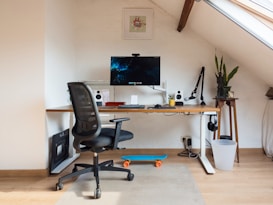 A modern, organized home office setup in an attic space features a sleek desk with a computer monitor, keyboard, and mouse. Surrounding the monitor are two small speakers, a microphone on an adjustable arm, and a potted plant. A black office chair is positioned at the desk. A skateboard rests on the floor nearby, and there is a small plant on a wooden stand beside the desk. A window in the sloped ceiling provides natural light.