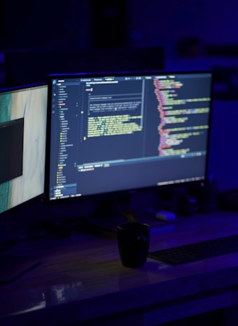 A dimly lit workspace featuring dual computer monitors displaying code editors with colorful syntax highlighting. A black coffee mug is placed on a wooden desk in front of the monitors, along with a keyboard. The overall setting suggests a programming or coding environment.