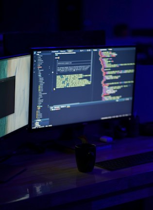 A dimly lit workspace featuring dual computer monitors displaying code editors with colorful syntax highlighting. A black coffee mug is placed on a wooden desk in front of the monitors, along with a keyboard. The overall setting suggests a programming or coding environment.