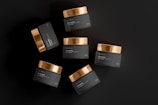 An arrangement of six dark cosmetic jars with gold lids spread out against a matte black background. The labels on the jars indicate they are for cosmetic or beauty products.