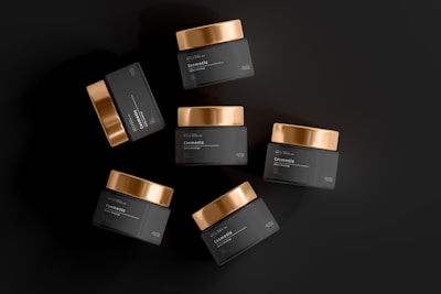 An arrangement of six dark cosmetic jars with gold lids spread out against a matte black background. The labels on the jars indicate they are for cosmetic or beauty products.