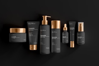 A collection of black and gold cosmetic containers arranged in a row. The set includes a jar, tubes, bottles with pumps, and a dropper bottle. Each item is designed with a matte black finish and shiny gold accents, giving a luxurious and elegant appearance. The products are labeled with the word 'cosmetic' and have simple, minimalistic typography.
