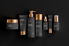 A collection of black and gold cosmetic containers arranged in a row. The set includes a jar, tubes, bottles with pumps, and a dropper bottle. Each item is designed with a matte black finish and shiny gold accents, giving a luxurious and elegant appearance. The products are labeled with the word 'cosmetic' and have simple, minimalistic typography.