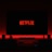 A television screen displays the Netflix logo in red against a black background. The room is dimly lit with a red glow possibly from LED backlighting behind the screen. Two speakers are positioned on either side of the TV set atop a minimalist entertainment center.