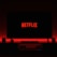 A television screen displays the Netflix logo in red against a black background. The room is dimly lit with a red glow possibly from LED backlighting behind the screen. Two speakers are positioned on either side of the TV set atop a minimalist entertainment center.