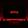 A television screen displays the Netflix logo in red against a black background. The room is dimly lit with a red glow possibly from LED backlighting behind the screen. Two speakers are positioned on either side of the TV set atop a minimalist entertainment center.
