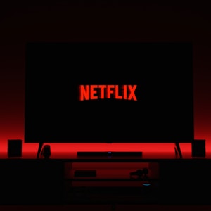 A television screen displays the Netflix logo in red against a black background. The room is dimly lit with a red glow possibly from LED backlighting behind the screen. Two speakers are positioned on either side of the TV set atop a minimalist entertainment center.